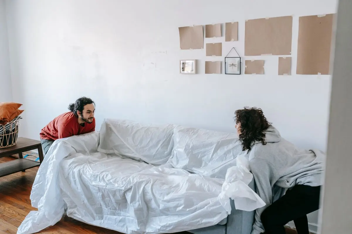 Two people lifting a sofa protected with a clear furniture cover.